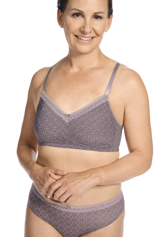 Woman wearing a matching lavender Amoena wire-free mastectomy bra and panty set, smiling confidently against a white background